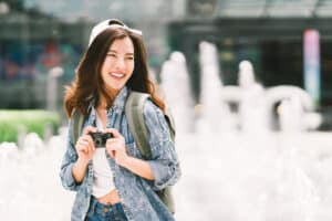 Young beautiful Asian backpack traveler woman using digital compact camera and smile, looking at copy space. Journey trip lifestyle, world travel explorer or Asia summer tourism concept