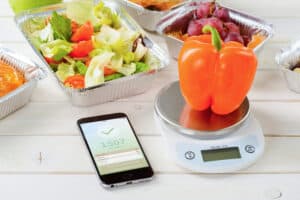 Calorie counter app on the smartphone, a kitchen scale and a fresh pepper on the wooden surface, close-up. Lettuce and tomato salad, grapes on the background. Counting calories.