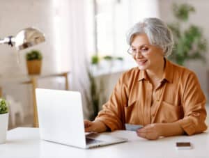 Cheerful elderly lady with credit card smiling and using laptop while sitting at table and doing online shopping at home