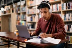 Young black student reading while using laptop and studying in a library.