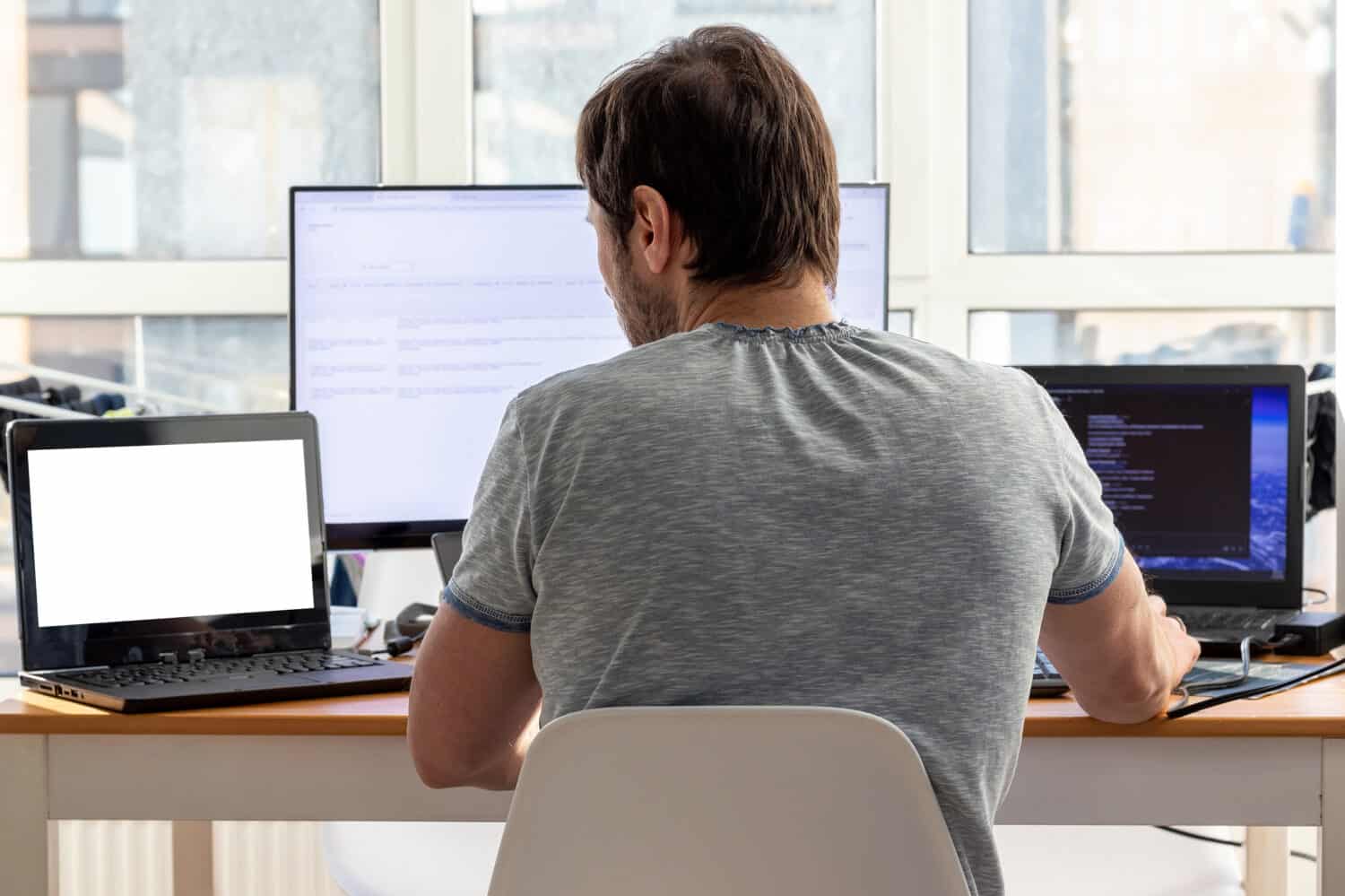 A man in a gray t-shirt is sitting at a workplace with two laptops and a monitor near the window.