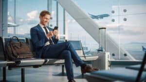 Airport Terminal Flight Wait: Smiling Businessman Uses Smartphone for e-Business, Browsing Internet with an App. Traveling Entrepreneur Work Online on Mobile Phone in Boarding Lounge of Airline Hub