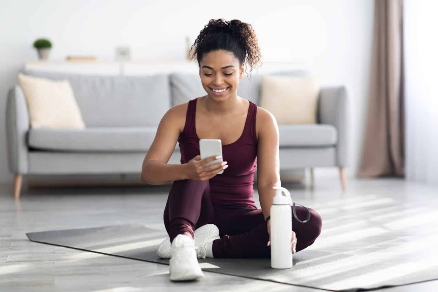 Woman in sportswear sitting on yoga mat at home, holding bottle of water and using the Down Dog yoga app