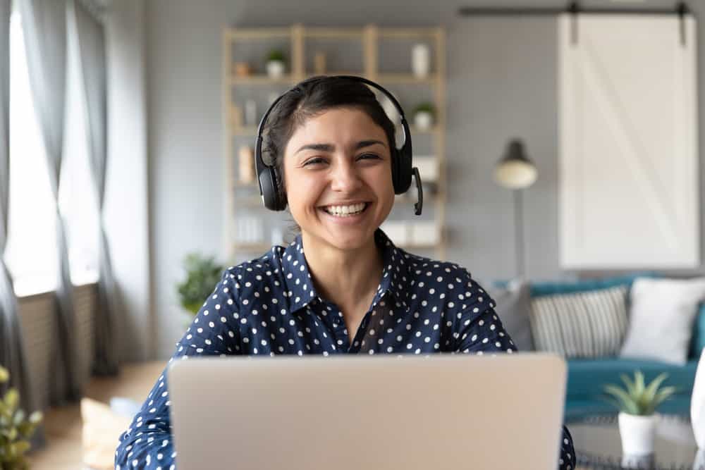 Head shot of a cheerful smiling IT Support Specialist sitting at table with computer, wearing headphones with mic, looking at camera.