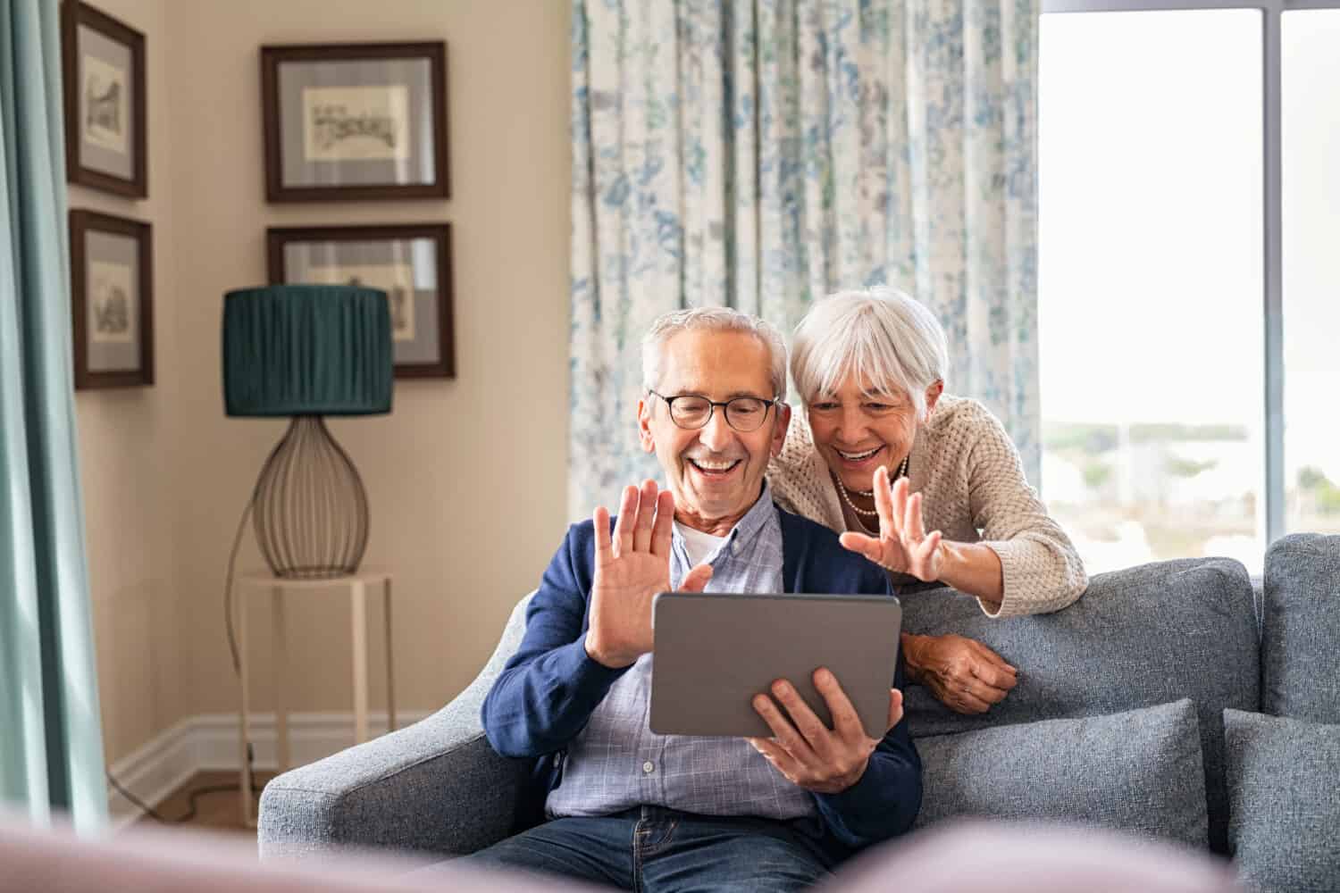 Happy senior couple using digital tablet for video conference and waving hand. Old man and smiling wife using digital tablet for video call with nephews or friends.