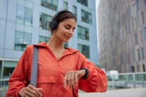 Female runner looks at smartwatch while going for run checks time listens music via headphones dressed in jacket strolls outdoor against modern tall buildings tracks wearable device wanders in city