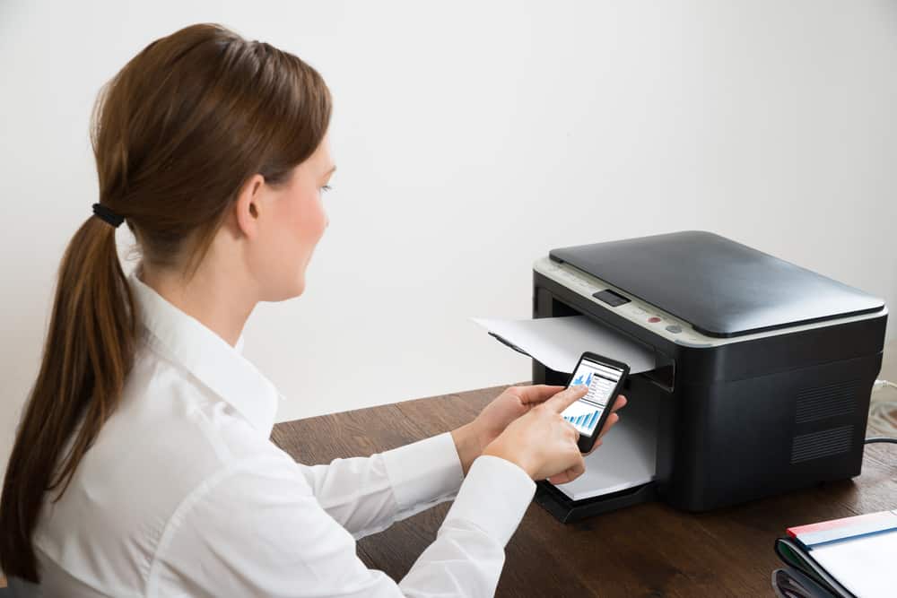 Young Businesswoman With Mobile Phone Showing Graph Connected To Printer At Desk