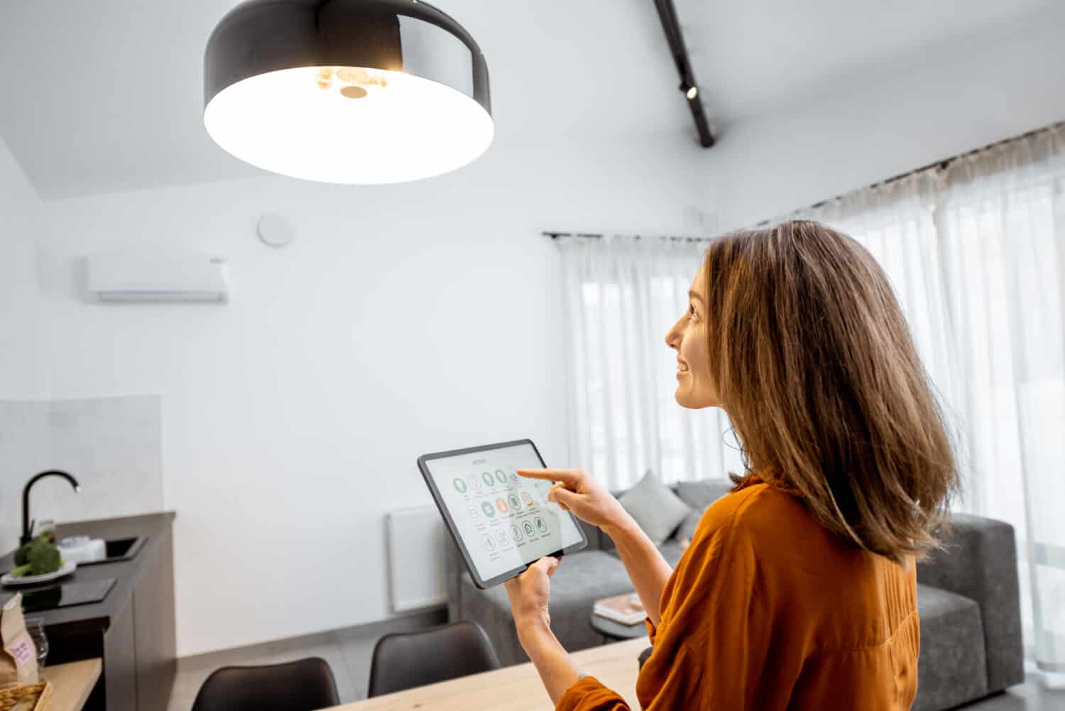 Young woman controlling home smart light with a digital tablet in the living room. Concept of a smart home and light control with mobile devices