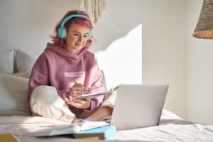 Hipster teen girl school student with pink hair wear headphone write notes watch video online webinar learn on laptop sit in bed distance elearning course video conference pc call in bedroom at home.