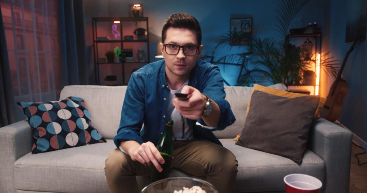 Close up of Caucasian young handsome guy in glasses watching television choosing channel while sitting on sofa and drinking beer from bottle. Man watch movie on TV sitting on couch at home, leisure