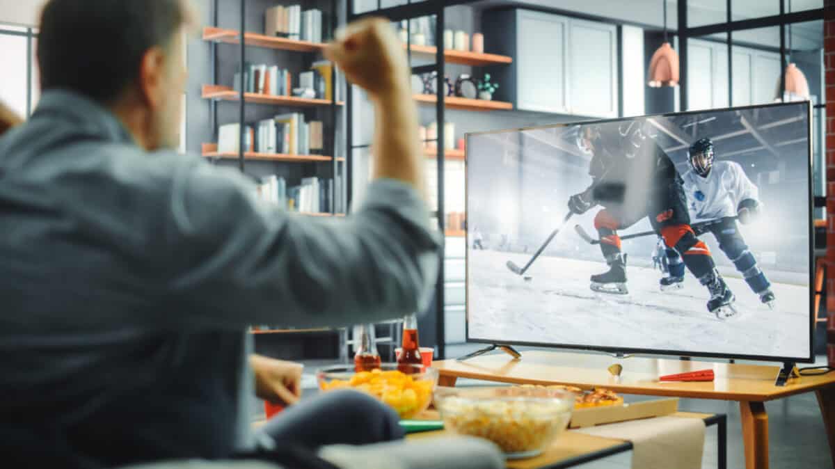 At Home Ice Hockey Fans Sitting on a Couch Watch Game on TV, Cheer when Favourtite Sports Team to Win the Championship. Screen Shows Professional Players During World Cup. Over the Shoulder