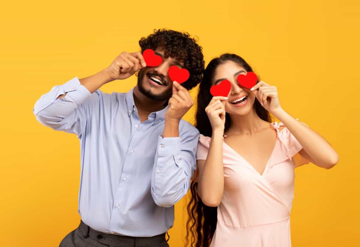 Lovers blinded by their big love. Young indian couple in love holding red heart-shaped cards over eyes and smiling, standing over yellow studio background. St. Valentines Day celebration concept
