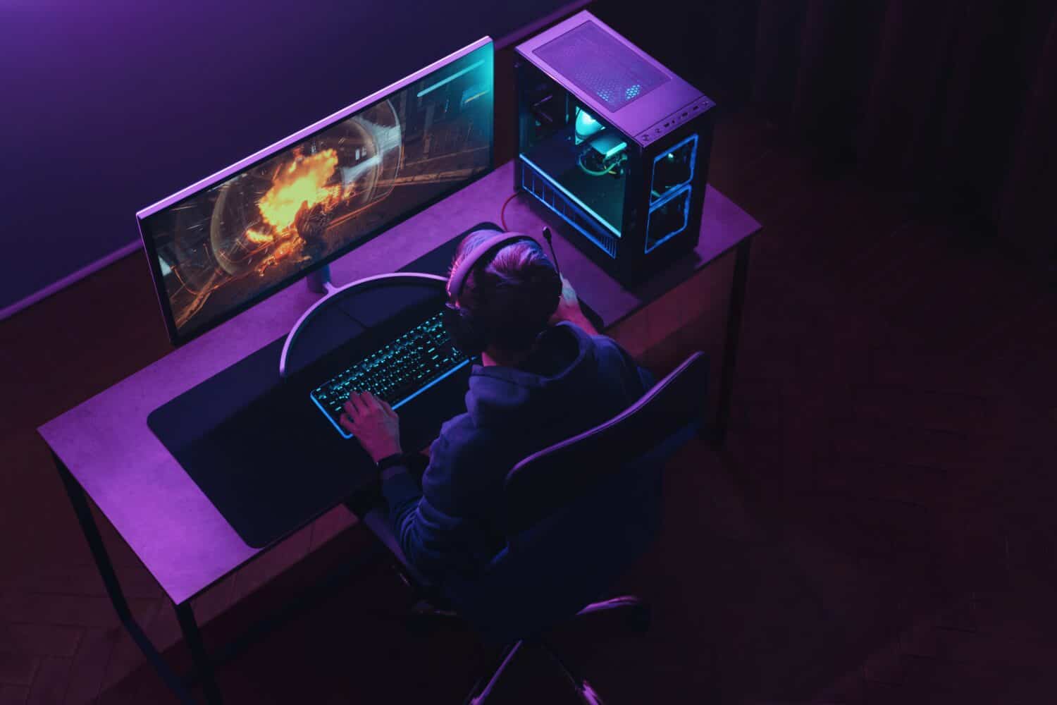 Top view of professional cyber sportsman in headphones playing shooter video game on his powerful gaming PC in dark neon room at night. Pro gamer participates in online esport tournament. Cyber sport