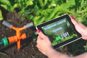 Woman is holding in hand a tablet computer with a program of irrigation control system on a garden sprinkler background.