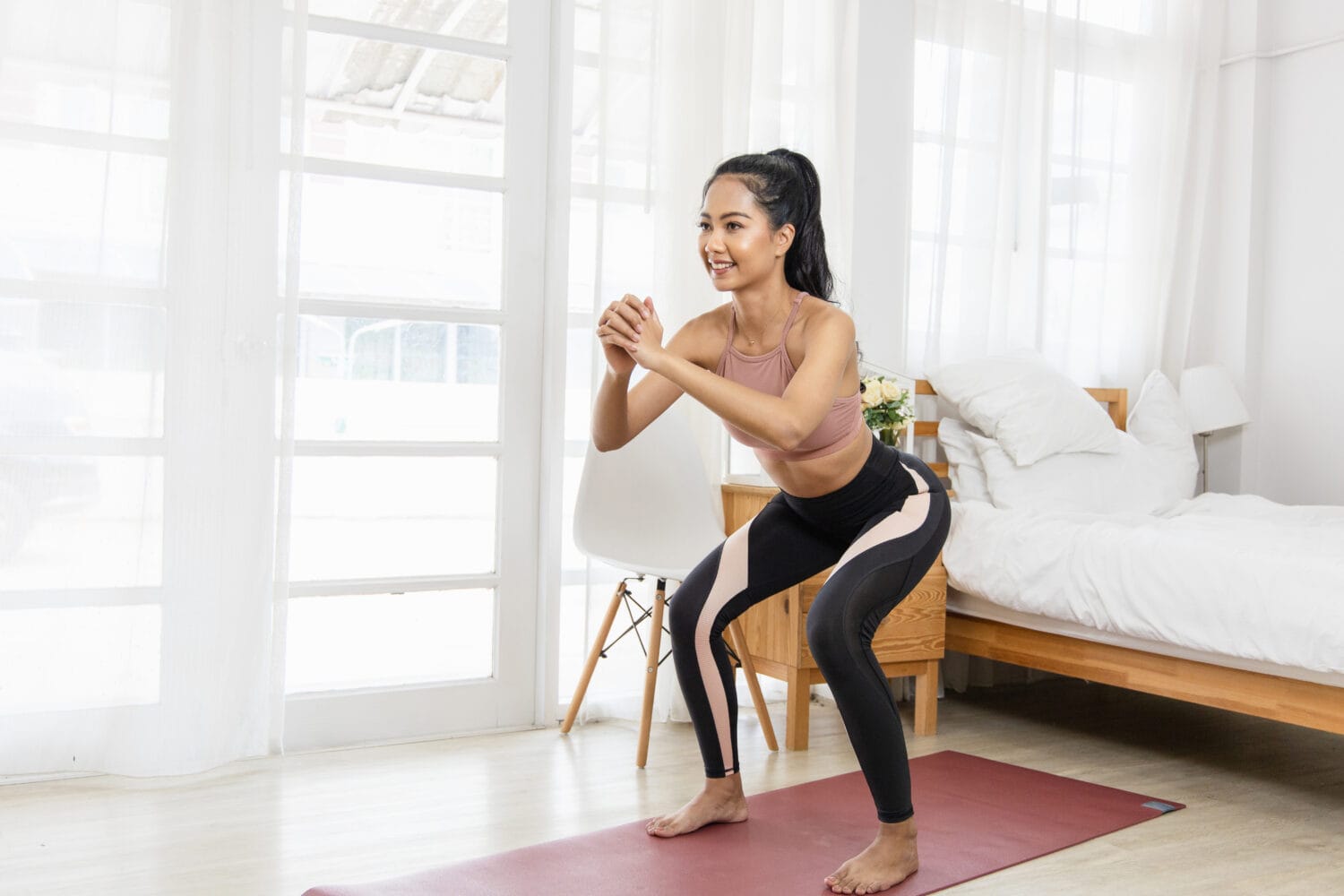 A beautiful Asian woman's fitness at home instead of going to the gym. She is doing squats on a yoga mat in the bedroom. She wears sportswear. Exercise concept for good shape