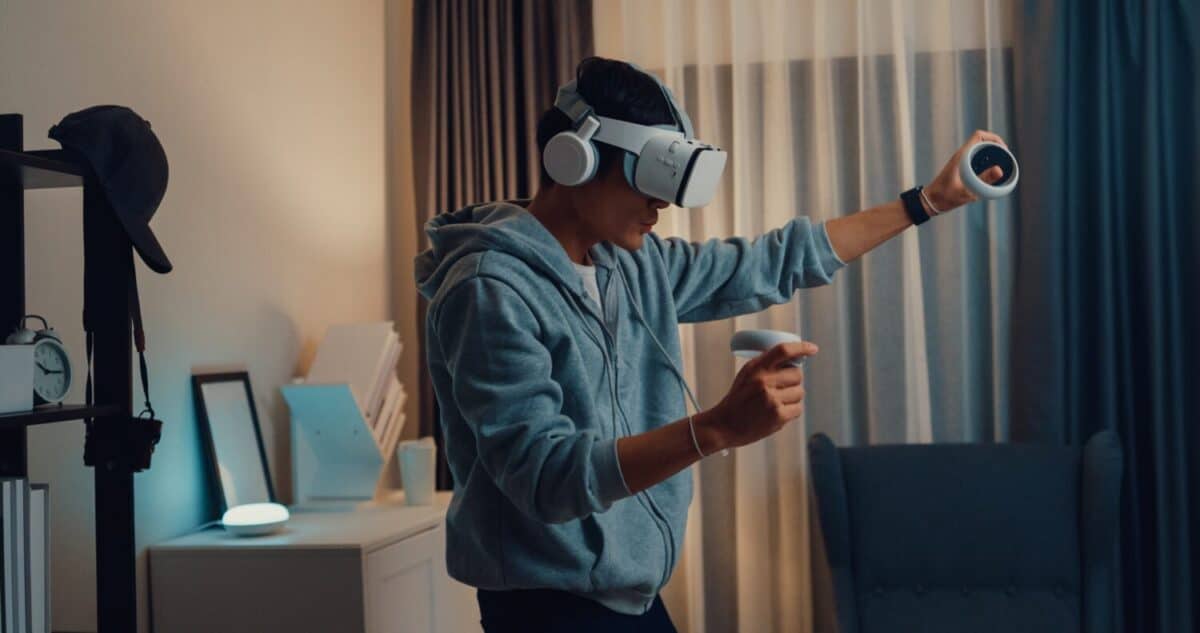 Young Asian man gamer wear gray hoodie with VR oculus hold VR joystick controllers feeling exciting with new experience of game stand front sofa living room home at night. Virtual reality concept.