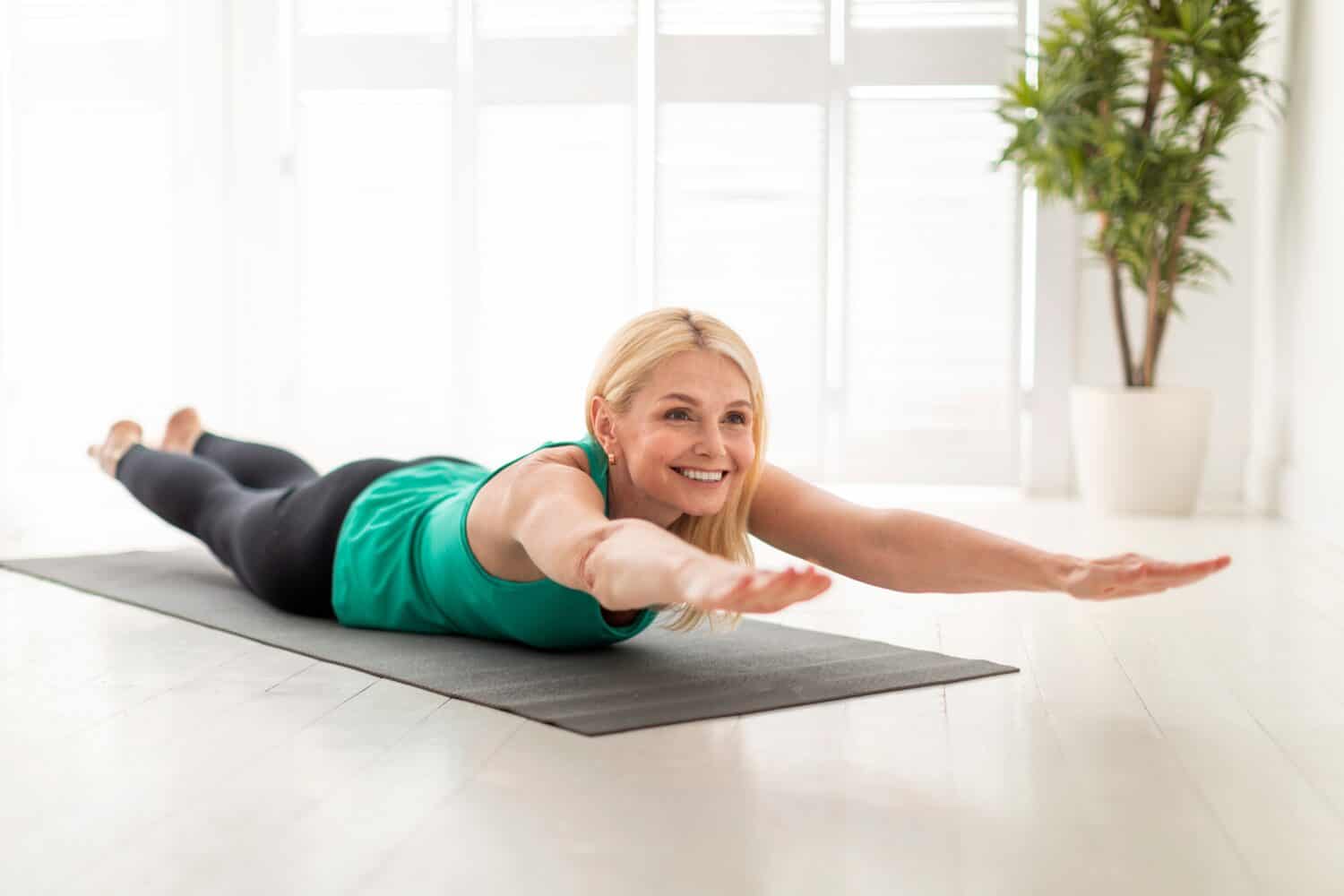Sporty Senior Woman Making Superman Exercise While Training At Home, Smiling Mature Lady Doing Back Bending While Lying On Fitness Mat In Light Living Room, Raising Arms And Legs, Free Space