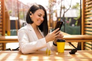 Brunette Caucasian business lady with small wireless black headphones in her ears looks at the phone with a smile, reading messages. Woman is relaxing in a cafe on the terrace enjoying music