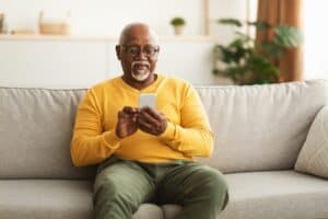 Senior Black Man Using Smartphone Texting Networking Online And Scrolling News In Social Media Application Sitting On Couch At Home. Mature Male Reading Message On Phone. New Mobile App Concept