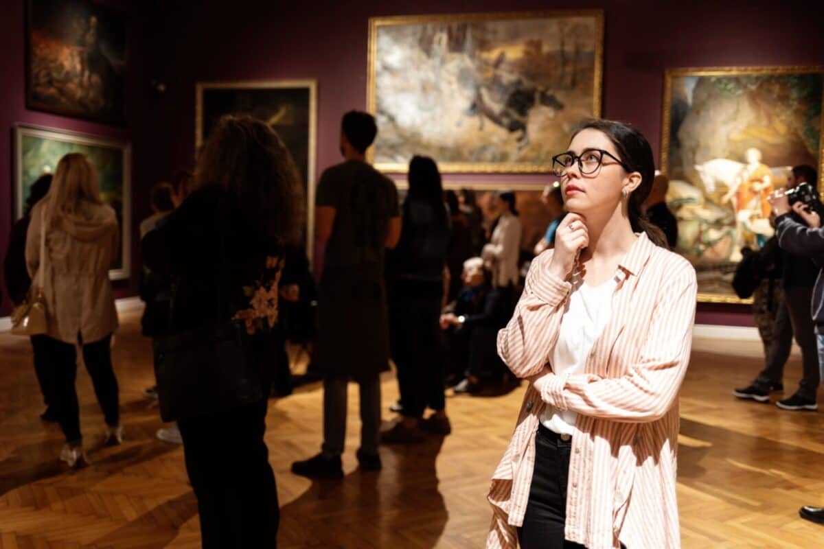 Thoughtful young Caucasian woman wearing glasses and looking at exhibition. In background, people are looking at paintings. Concept of Museum Day