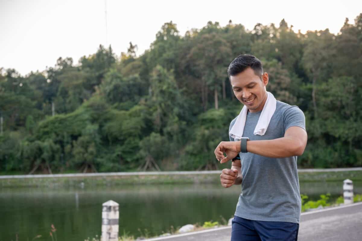 A healthy, happy, and fit Asian man in sportswear is checking his running miles and calories burning on his smartwatch while running outdoors. Exercise and modern gadget concepts