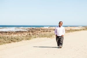 dwarfish african man strolling happily down the dirt road on a rocky coast wearing a smart-casual attire