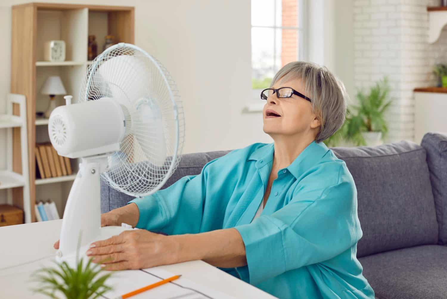 Senior woman, exhausted from sweltering summer heat, uses electric fan to freshen up. Old grandma, tired of extreme heatwave, enjoying fresh air while sitting on couch by table with ventilator at home