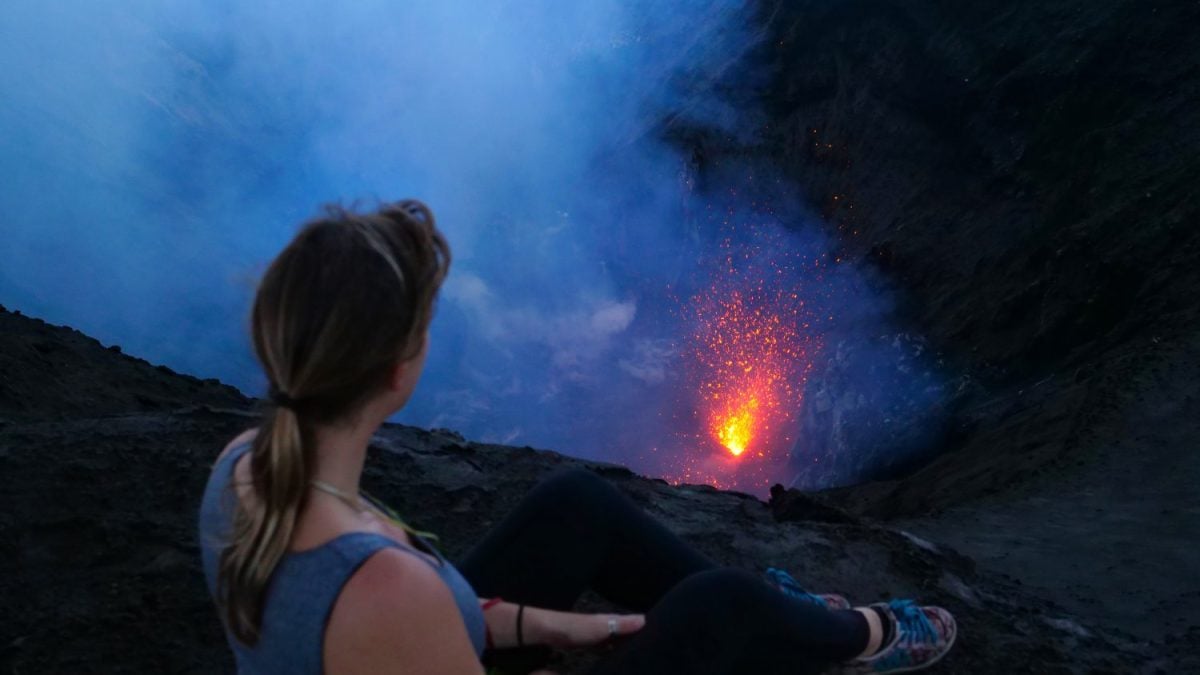 CLOSE UP: Young traveler woman sits on the crater rim of stunning Mount Yasur in Vanuatu. Unrecognizable female tourist observing the smoke and ash rising from the active volcano roaring below her.