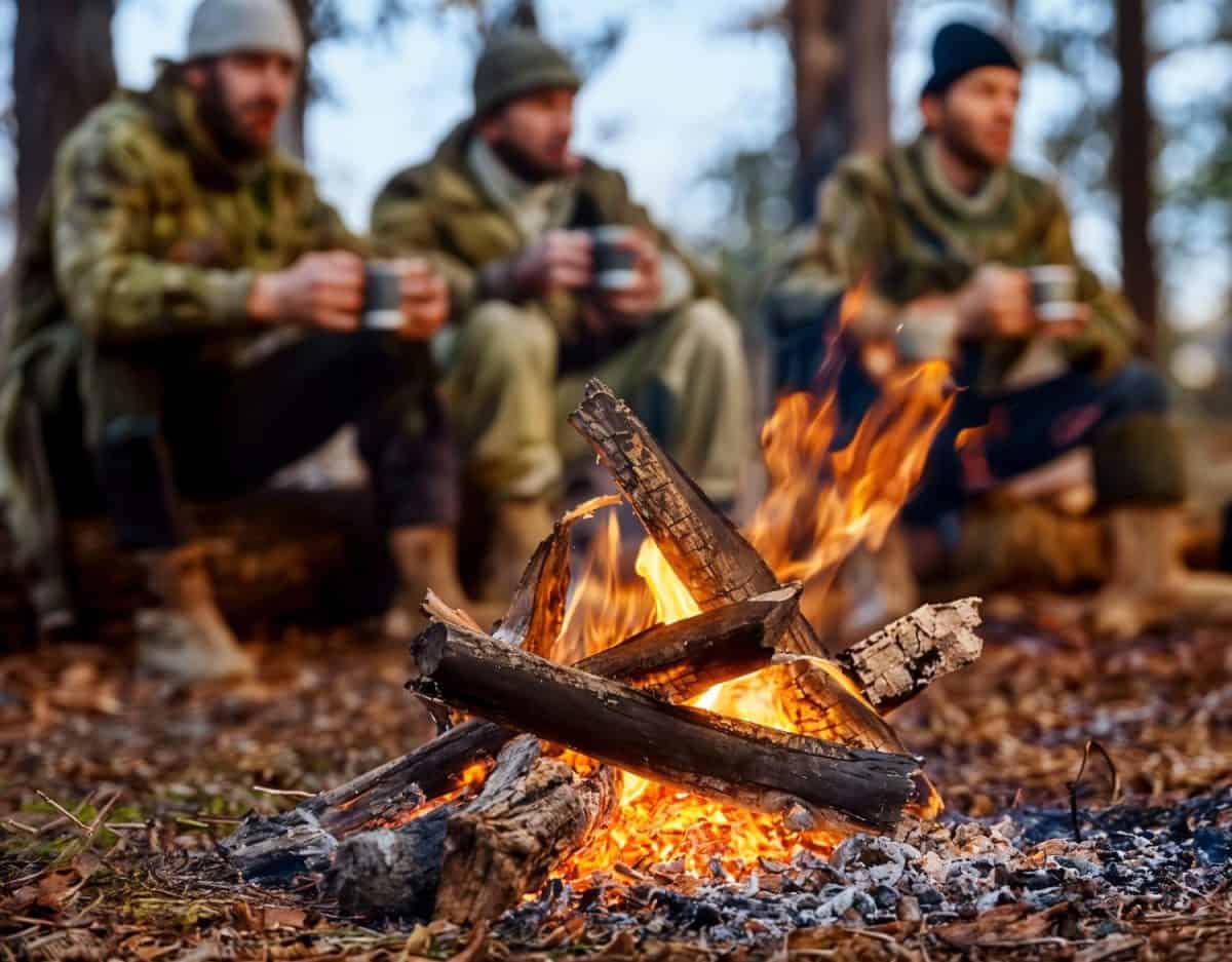 Three hunters in winter gear sit around a campfire in the forest, enjoying the warmth and hot drinks after a day of hunting