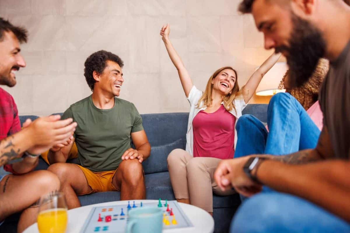 Group of cheerful young friends having fun playing ludo board game while spending time together at home
