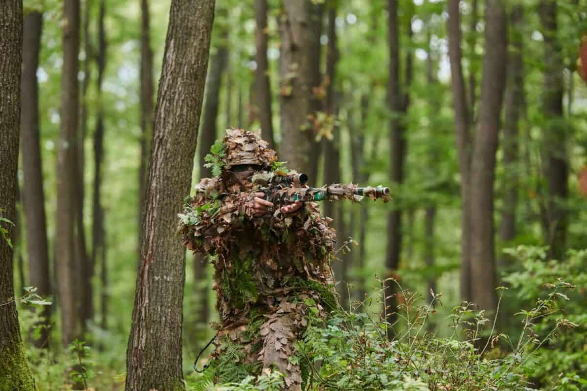 A highly skilled elite sniper, camouflaged in the dense forest, stealthily maneuvers through dangerous woodland terrain on a covert and precise mission