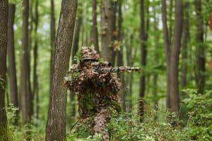 A highly skilled elite sniper, camouflaged in the dense forest, stealthily maneuvers through dangerous woodland terrain on a covert and precise mission